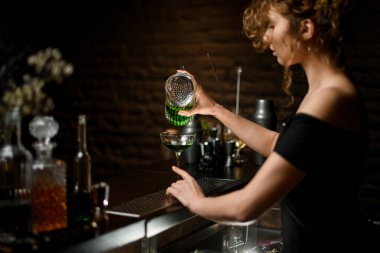 Koyu bardaki profesyonel kadın barmen bardaktan bardağa kokteyl doldurur.