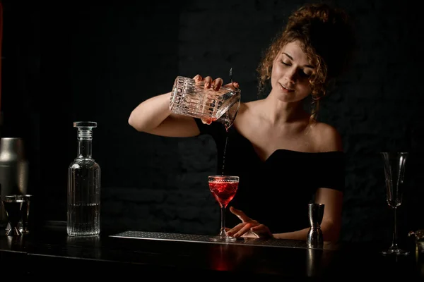 Smiling woman bartender in dark bar pours cocktail from cup to glass — Stock Photo, Image