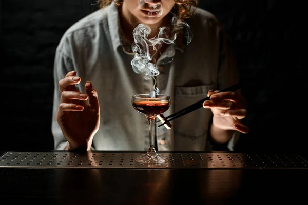 Молодая женщина бармен держит пинцет с дымящейся палкой возле стекла — стоковое фото