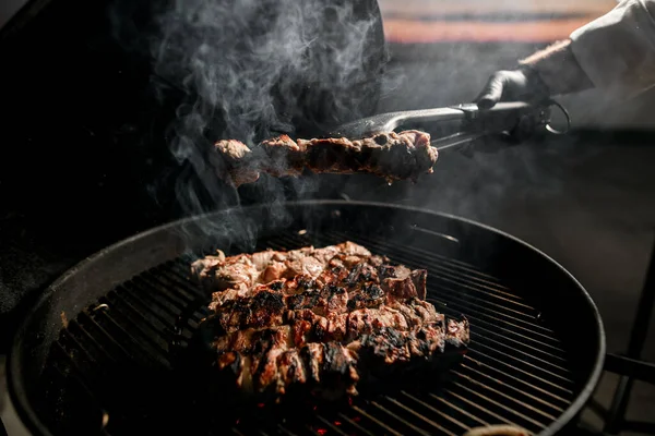 Masculino chefs mão usando pinças detém pedaço de carne sobre churrasco . — Fotografia de Stock