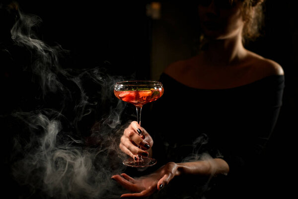 Young beautiful woman holds glass with bright cocktail in front of her at dark bar.