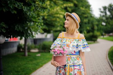 Süslü çiçek kutusuyla sarışın bir kadın parkta yürüyor.