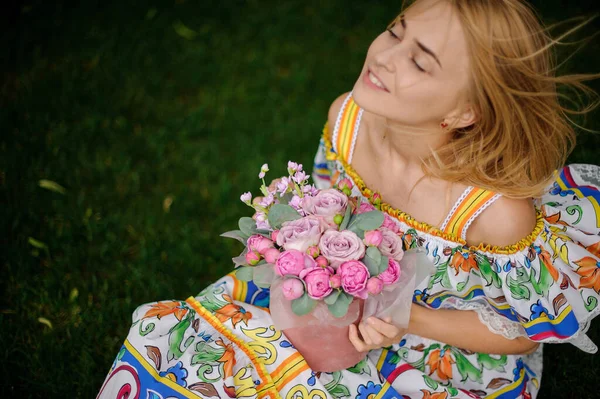 Усміхнена блондинка з закритими очима сидить на газоні і тримає круглу коробку з трояндами — стокове фото