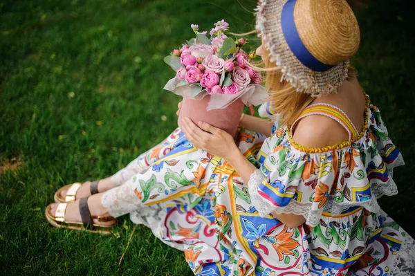 Молода жінка в капелюсі і барвисте плаття сидить на галявині з букетом — стокове фото