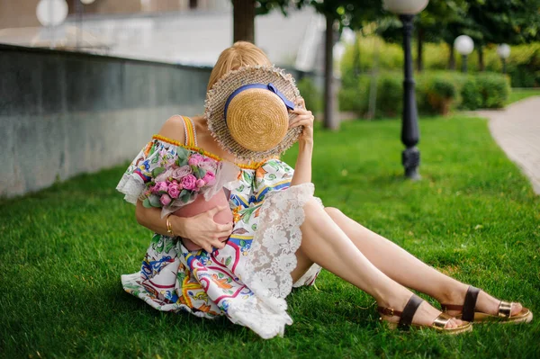 Young woman sits on lawn with bouquet and covers her face with hat — Stock Photo, Image