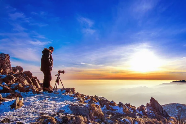 Fotógrafo profesional toma fotos con cámara en trípode en pico rocoso al atardecer . — Foto de Stock