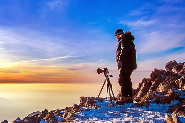 专业摄影师带照片在岩石峰上三脚架上的相机在日落时分. — 图库照片