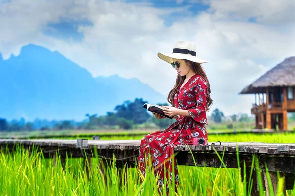 Mujer joven leyendo un libro y sentada en un camino de madera con campo de arroz verde en Vang Vieng, Laos . —  Fotos de Stock