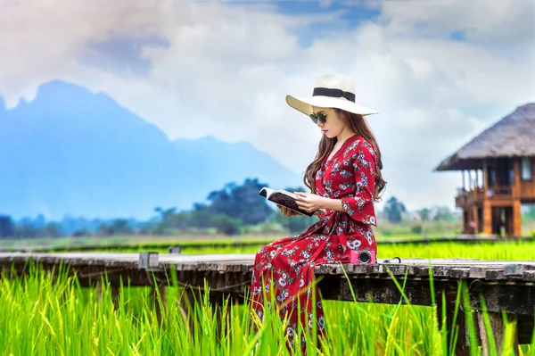 Mujer joven leyendo un libro y sentada en un camino de madera con campo de arroz verde en Vang Vieng, Laos . —  Fotos de Stock