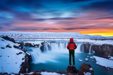 Kışın gün batımında İzlanda 'da Godafoss şelalesi. Kırmızı ceketli adam Godafoss şelalesine bakıyor..