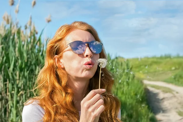 Menina de cabelos vermelhos em óculos de sol sopra dente-de-leão — Fotografia de Stock