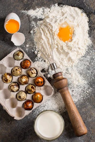 Baking ingredients. Flour, eggs, milk in the glass, quail eggs, eggbeater on a black stone texture from above. Flat lay. Rustic style.