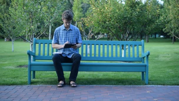 Сидящий на скамейке в парке мужчина, работающий на планшете — стоковое видео