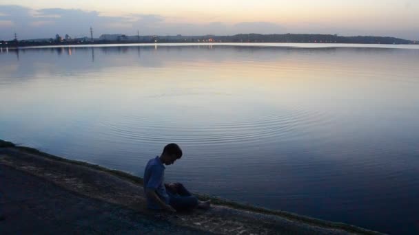 Молодий стрункий чоловік кидає камені на воду ввечері — стокове відео