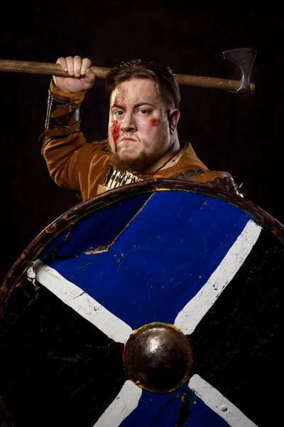 A Scandinavian warrior viking wounded in leather and metal armor with a fighting axe on a dark background. Blood and chopped wound