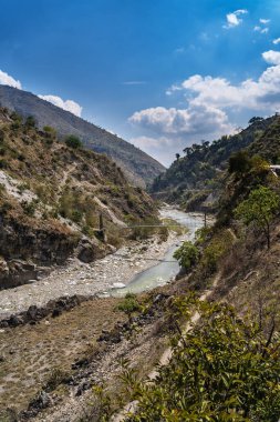 Himalayalar 'da bir dağ nehrinden geçen metal bir asma köprü. Güneşli bir gün, alçak dağlar dar vadiyi çevreleyen görünür..