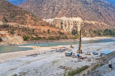 Nepal 'in vadilerinden birindeki bir dağ nehri üzerindeki köprünün sütunlarını beton ile doldurmak için hazırlık çalışmaları sürüyor. Görünür çalışma ekipmanları - kazıcı ve kazıcı.