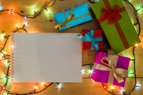 Presente cajas en el concepto de vacaciones de tarjetas de felicitación de fondo de madera. Navidad coloridas cajas de regalo en la mesa de madera. Marco de luces románticas de Navidad sobre fondo de madera con cuaderno abierto —  Fotos de Stock