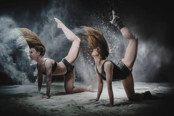 Two girls dance with flour in studio on black background, lights behind them and people helped girls