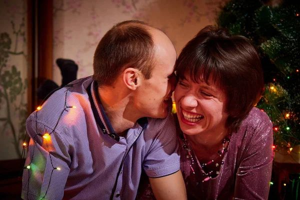 Adult couple staying at home together on holiday evening and lying on floor in front of christmas tree and hugging. Cute romantic lovers in New Year