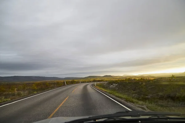 View on grey road, yellow surise and dramatic clouds from the car front window in early mourning. Driving car during sunshine in the tundra in Norway. Background of nature landscape and a road