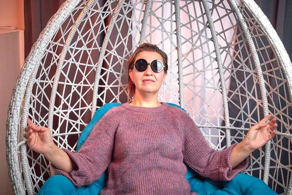 Ugly aged woman in a sweater and black glasses in a white wicker hanging chair in the room with curtains