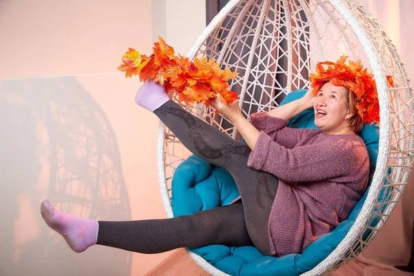 Ugly aged woman in a sweater with a wreath of yellow autumn maple leaves in a white wicker hanging chair in the room with curtains