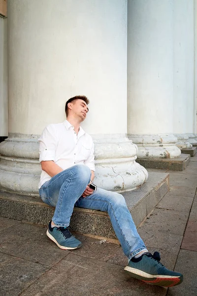 Young handsome man in jeans and a white shirt sitting near building with white columns in the city. The model pretends to be a bum and has a rest