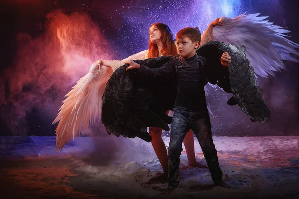 Black and white angel together on a dark background with colored lighting. The concept of war between good and evil. Girl and boy with angel wings during a photo shoot with flour and loose powder