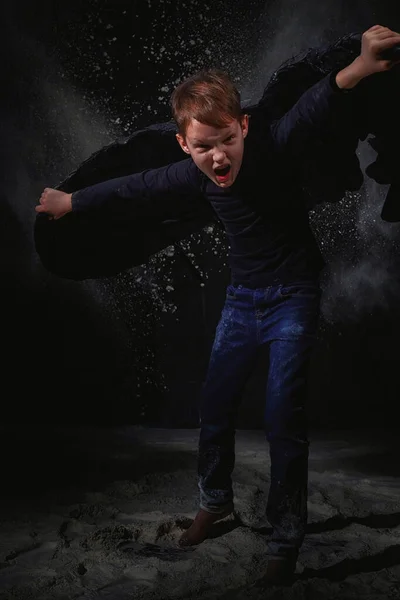 Black evil angel on a dark background with colored lighting. The concept of war between good and evil. Boy with angel wings during a photoshoot with flour and loose powder