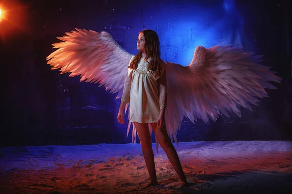 White angel on a dark background with colored lighting. The concept of war between good and evil. Girl with angel wings during a photoshoot with flour and loose powder