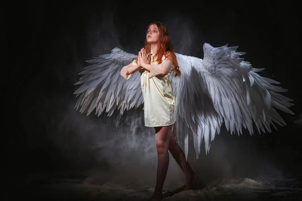 White angel on a dark background with colored lighting. The concept of war between good and evil. Girl with angel wings during a photoshoot with flour and loose powder