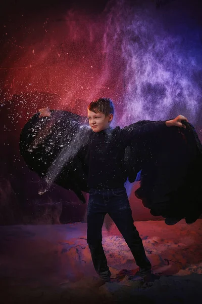 Black evil angel on a dark background with colored lighting. The concept of war between good and evil. Boy with angel wings during a photoshoot with flour and loose powder