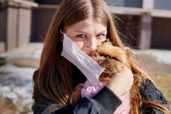 Girl trying to protect small dog from a coronavirus with a medical gauze white mask on the street in sunny spring day. Protection against disease during epidemics and pandemics. Covid-19 in Russia