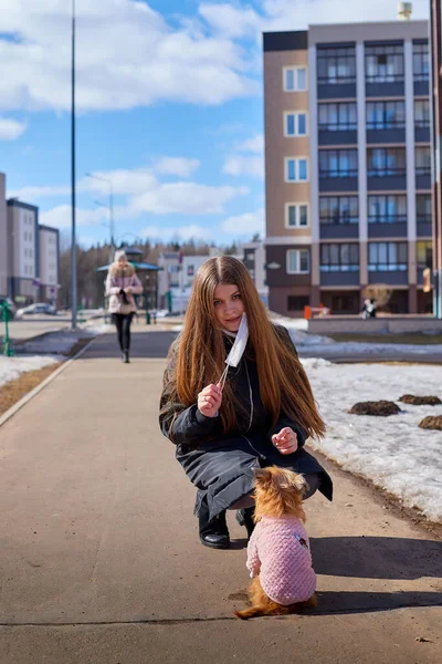 Girl in white medical gauze mask walking on the street with a dog in sunny spring day. Protection against disease and coronavirus during epidemics and pandemics. Covid-19 in Russia and russian city