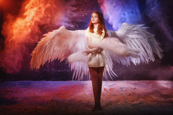 White angel on a dark background with colored lighting. The concept of war between good and evil. Girl with angel wings during a photoshoot with flour and loose powder