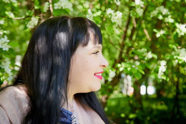 Portrait of fat chubby plump girl with black hair and blossoming apple tree with white flower background in the park in a spring day
