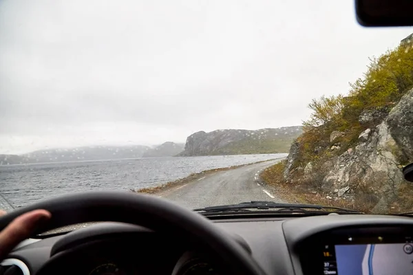 View from the car window of a sad, harsh, dramatic landscape with a lake, river or fjord, a rock in the distance on the horizon and a cloudy, rainy, gray sky