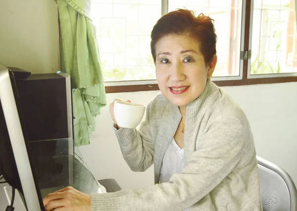 Asiática anciana mujer disfrutar de su café mientras se trabaja con el ordenador personal —  Fotos de Stock