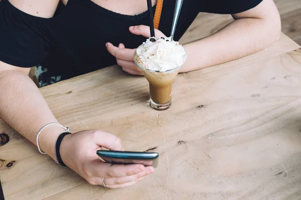 Girl drinking ice coffe and texting on her phone sitting on wood — Stock Photo, Image