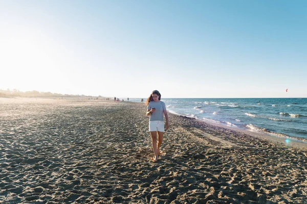 Bella ragazza che cammina sulla spiaggia concentrandosi sulla lettura sul telefono cellulare con retroilluminazione — Foto Stock