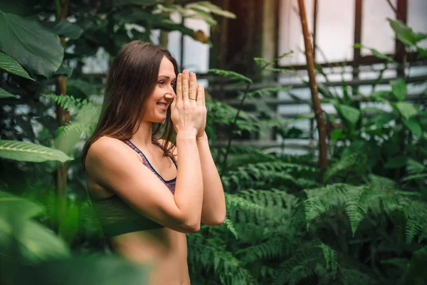 Young smiling yoga instructor doing namaste hand gesture at greenhouse — Stock Photo, Image
