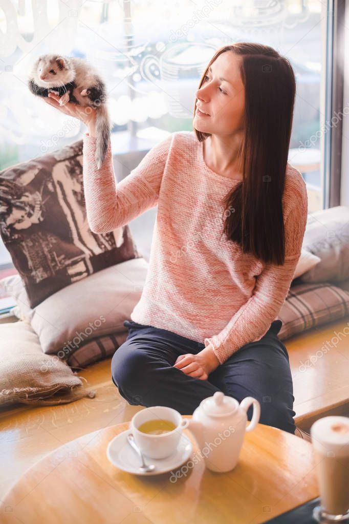 Happy young woman holding high her pet ferret in cafe