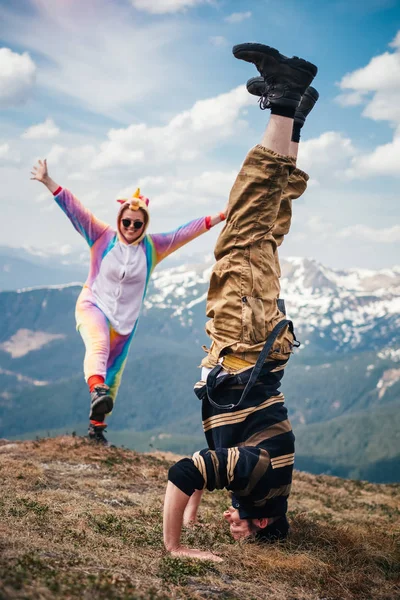 Funny travel concept, male hiker doing headstand while female clad in unicorn costume posing on his background high in mountains on a Spring day