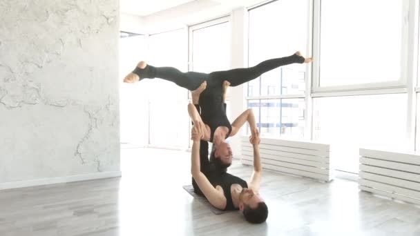 美丽的年轻女子练习杂技瑜伽练习与男子合作伙伴在白色健身工作室与大窗户的背景 — 图库视频影像
