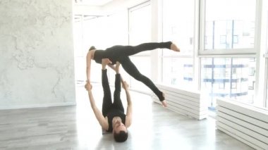 Arka planda büyük pencereler ile beyaz fitness stüdyosunda erkek ortağı ile güzel genç kadın uygulama akrostiş yoga egzersiz