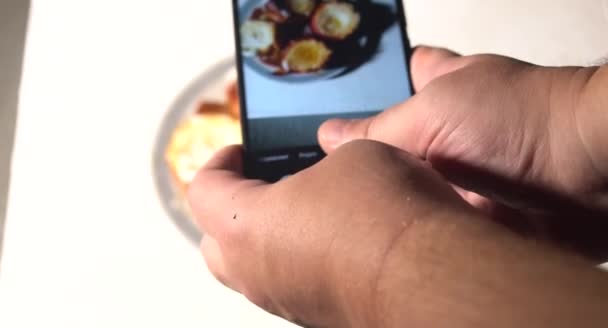 若い男が電話でレストランで昼食を撮っている。スマホでケーキの食べ物を写真に撮り、携帯カメラで食事を撮る青年. — ストック動画