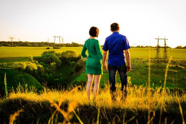 Kız ve erkek el ele tutuşup yeşil bir tepede duruyorlar. Gün batımı. Arka plan.