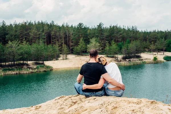 Дівчина і хлопець тримаються за руки і сидять на березі лісового озера. прекрасний вид на озеро. вид ззаду . — стокове фото