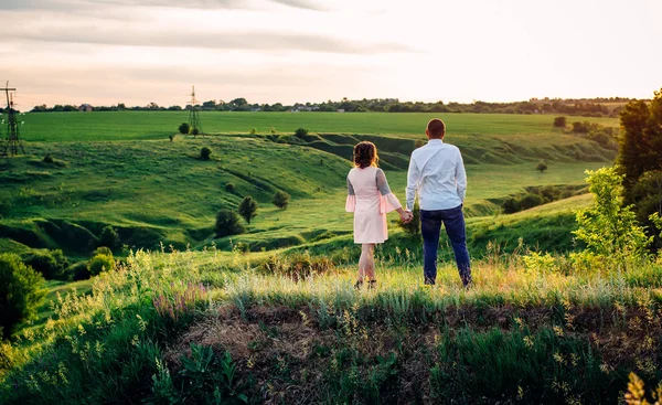 Meisje en jongen die elkaars hand vasthouden en op een groene heuvel staan. Zonsondergang. Achteraanzicht. — Stockfoto